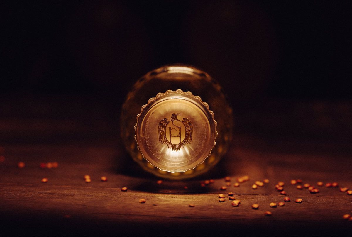 Vue 2 frontale en ombre et lumière sur le bouchon gravé du sceau Sainte Harmonie d'une bouteille couchée, entourée de grains de sorgho dispersés sur du bois — Sainte Harmonie, Baijiu Français.