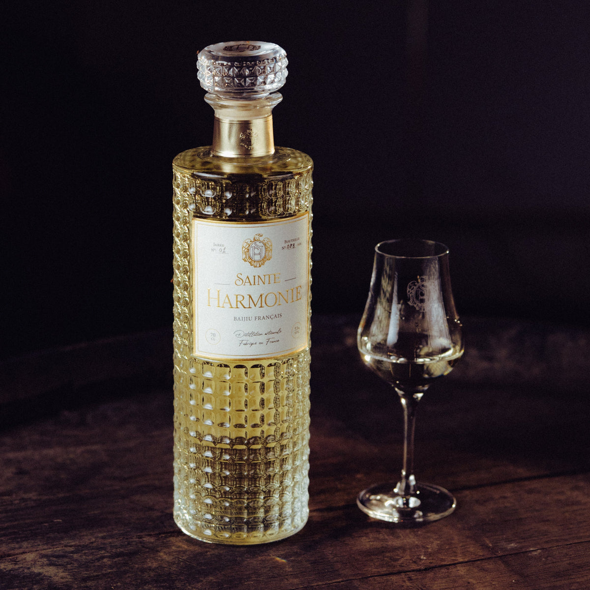 Flacon en verre ciselé d'exception Sainte Harmonie pose à côté d'un verre à dégustation tulipe sur une table en bois rustique dans une ambiance sombre et élégante - Sainte Harmonie, Baijiu Français.