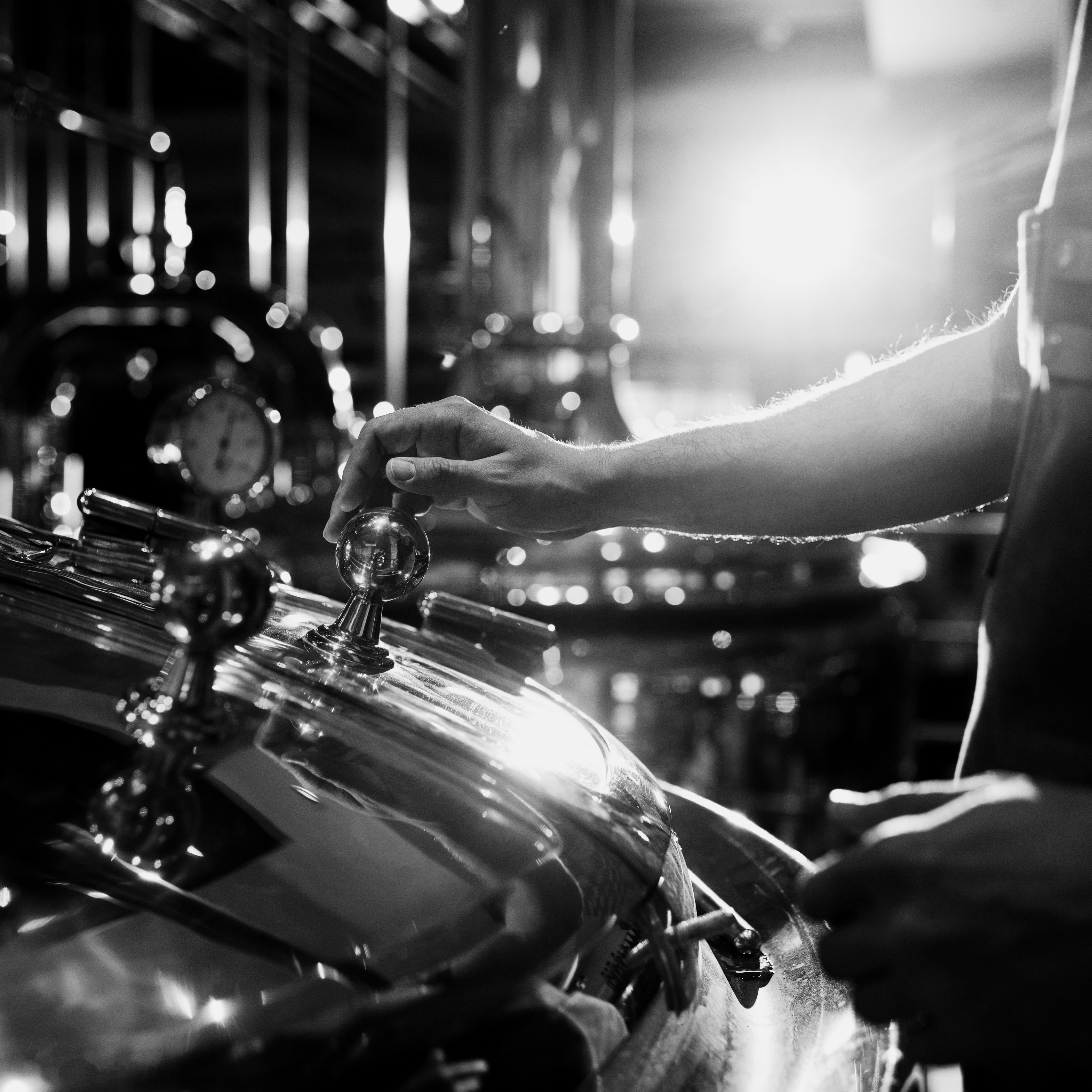 Photographie en noir et blanc de la main d'un maître distillateur ajustant les commandes d'un alambic en cuivre rutilant au cours de la distillation - Sainte Harmonie, Baijiu Français.