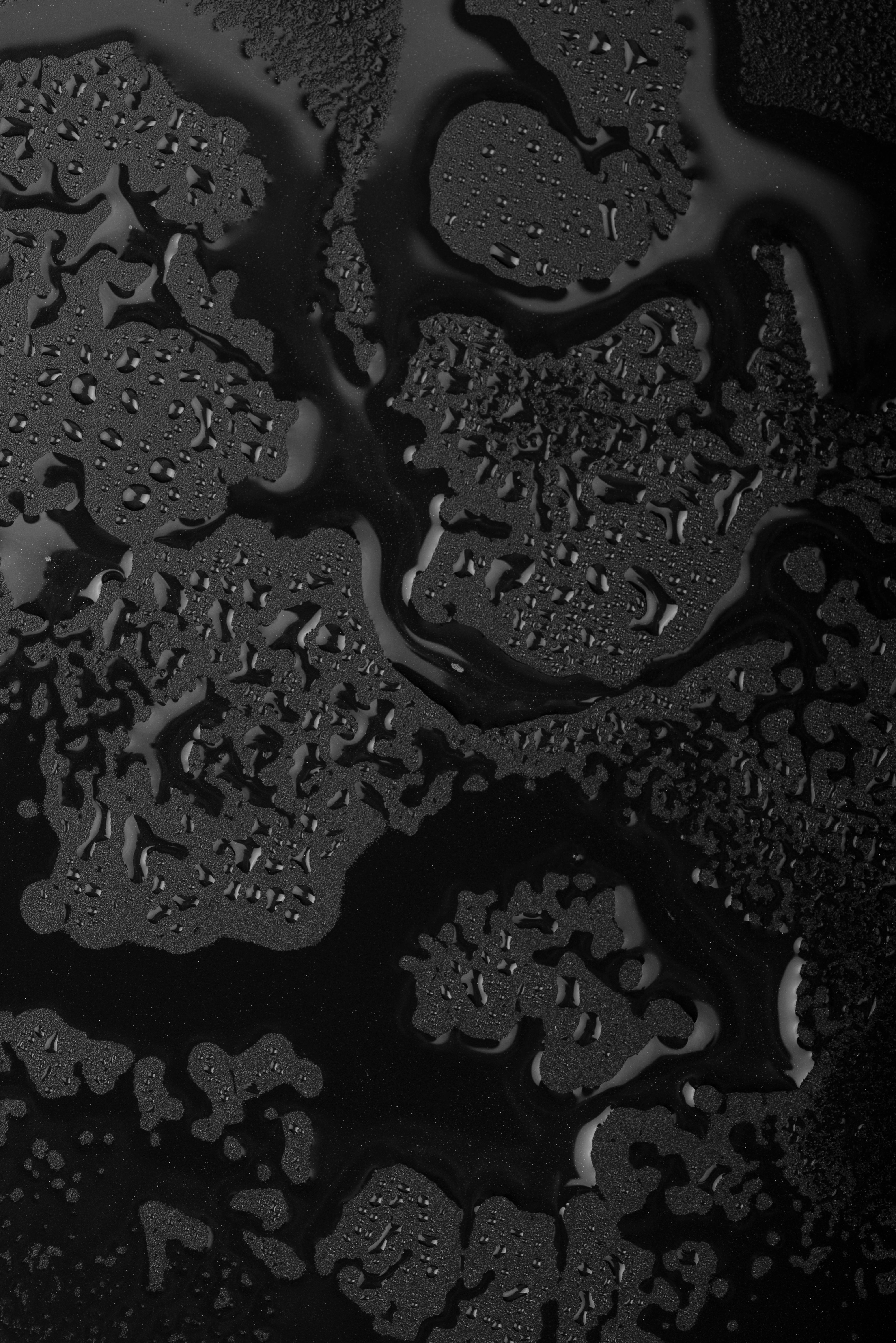 Vue zénithale de gouttelettes d'eau et de zones de condensation formant des motifs organiques abstraits sur une surface texturée noire et mate - Sainte Harmonie, Baijiu Français.