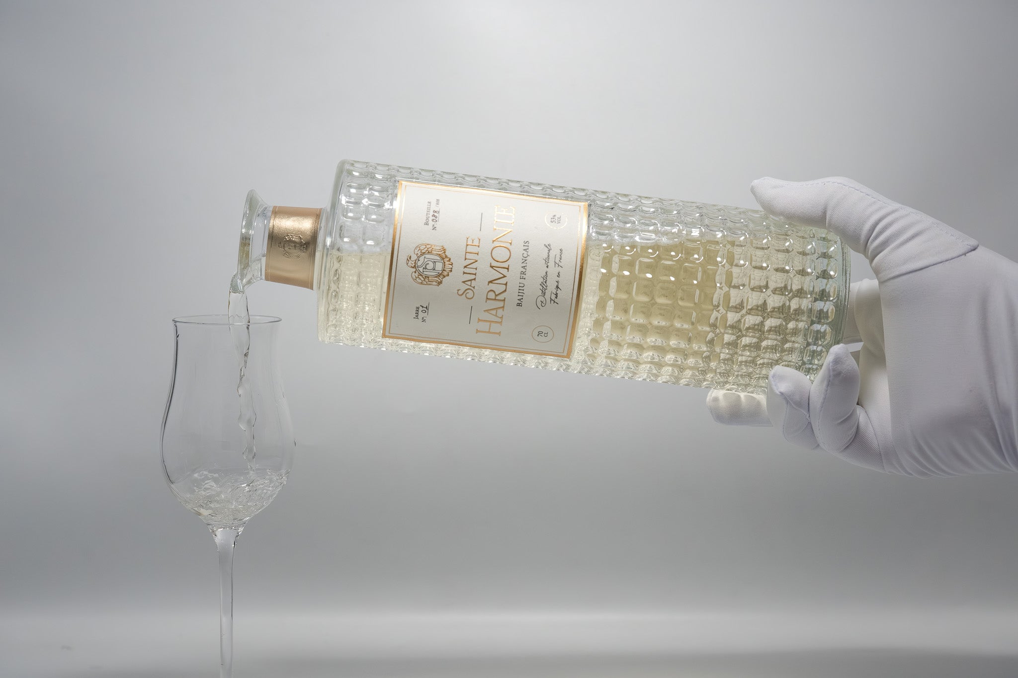 Main portant un gant blanc élégant servant délicatement le spiritueux d'un flacon en verre ciselé vers un verre à dégustation sur fond blanc épuré - Sainte Harmonie, Baijiu Français.