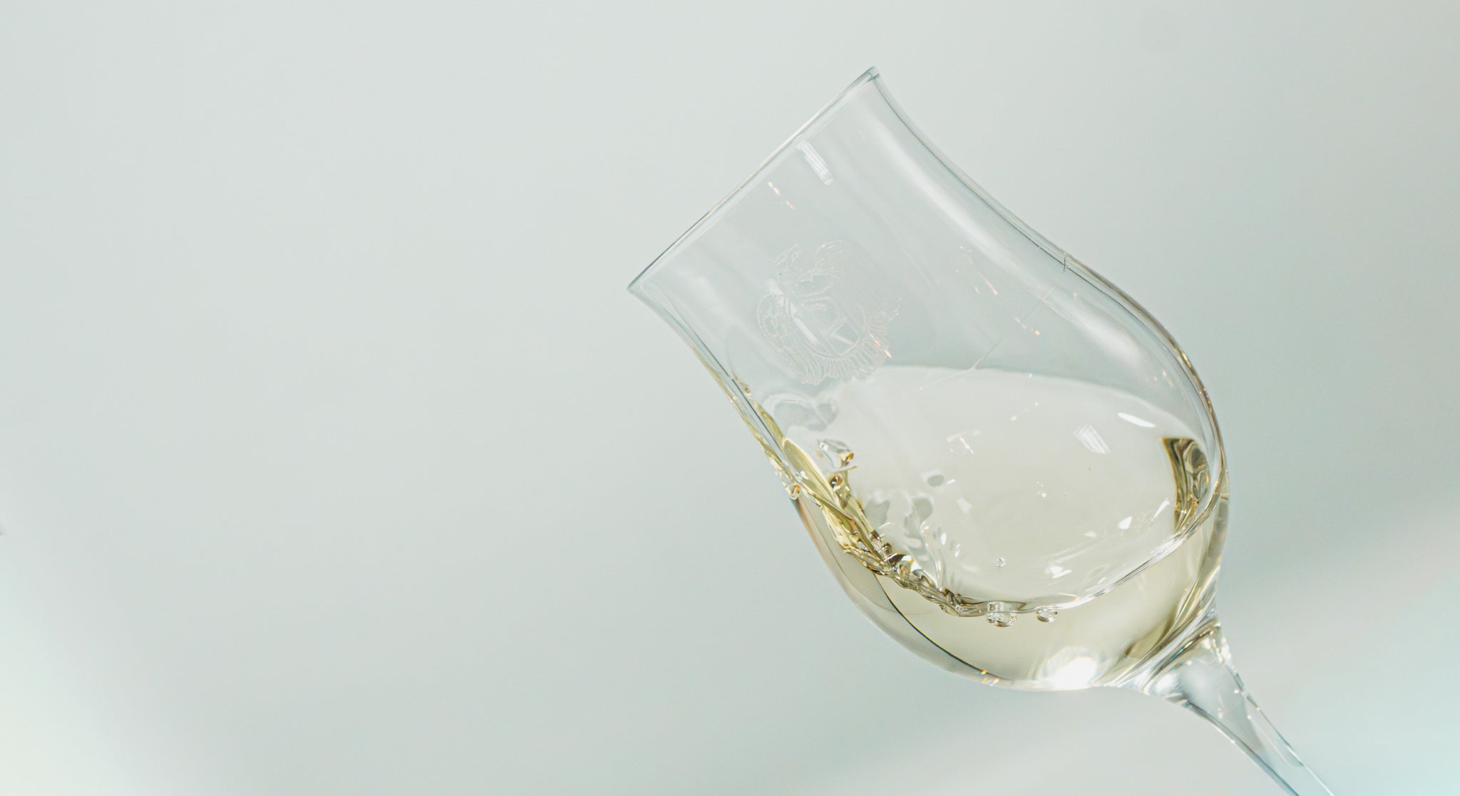 Gros plan incliné sur un verre tulipe gravé du logo de la marque, en plein mouvement d'aération pour sublimer la robe et les arômes du spiritueux blanc - Sainte Harmonie, Baijiu Français.