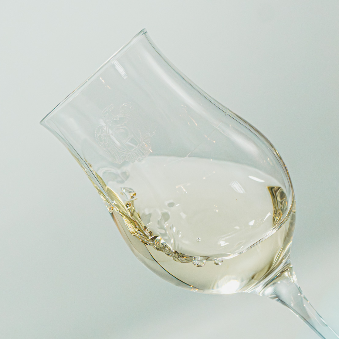 Gros plan sur un verre à dégustation gravé en mouvement, créant une ondulation du spiritueux blanc et limpide sur un fond clair épuré - Sainte Harmonie, Baijiu Français.