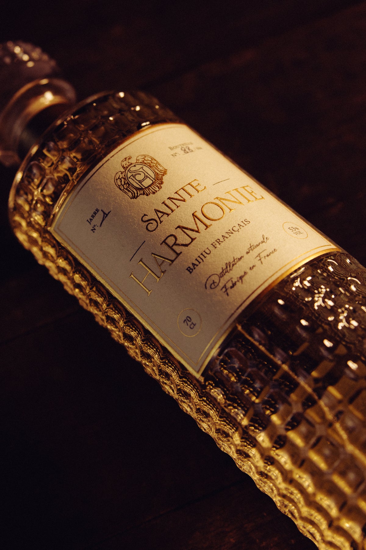 Gros plan dynamique en diagonale sur l'étiquette dorée et le travail du verre ciselé du flacon, capturant les reflets chauds et somptueux du spiritueux - Sainte Harmonie, Baijiu Français.