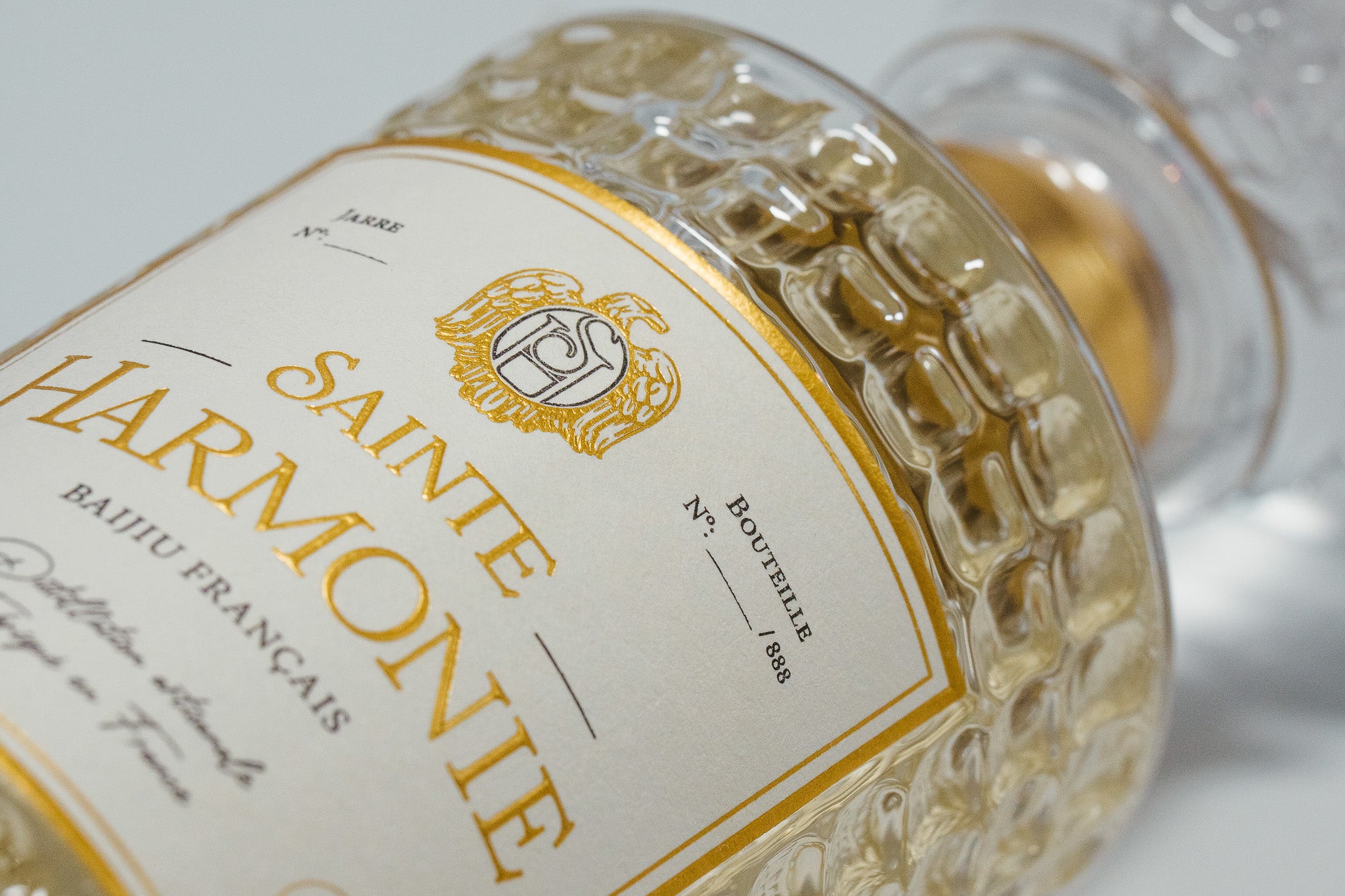 Gros plan sur l'étiquette texturée aux dorures à chaud et le verre ciselé du flacon d'édition limitée numérotée sur 888 - Sainte Harmonie, Baijiu Français.