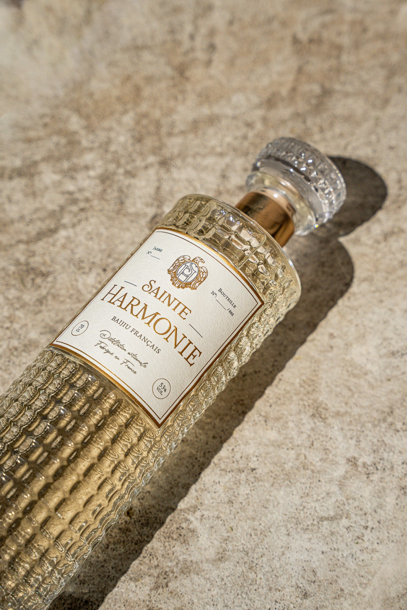Vue en contre-plongée et en diagonale de la bouteille de prestige Sainte Harmonie posée sur une table en pierre texturée sous une lumière naturelle ombragée - Sainte Harmonie, Baijiu Français.