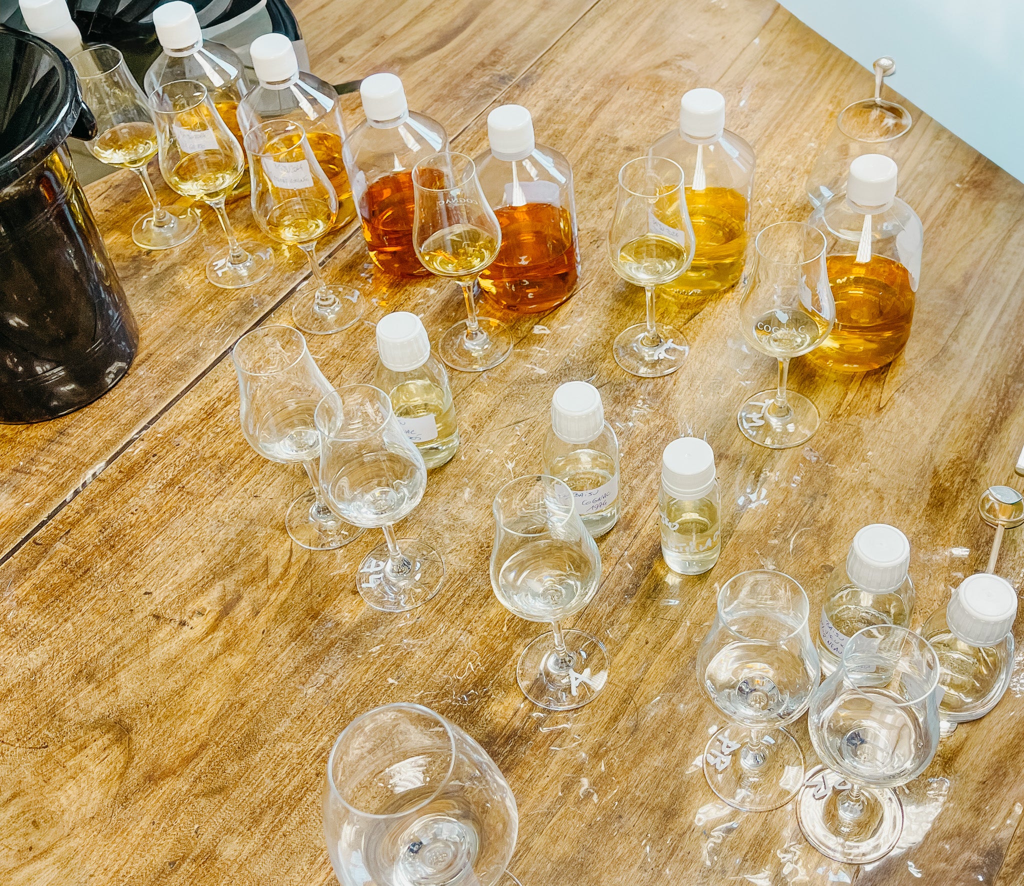 Vue surélevée d'une table de travail en bois partagée entre plusieurs verres de dégustation tulipe et des échantillons de spiritueux transparents et ambrés pour l'assemblage - Sainte Harmonie, Baijiu Français.
