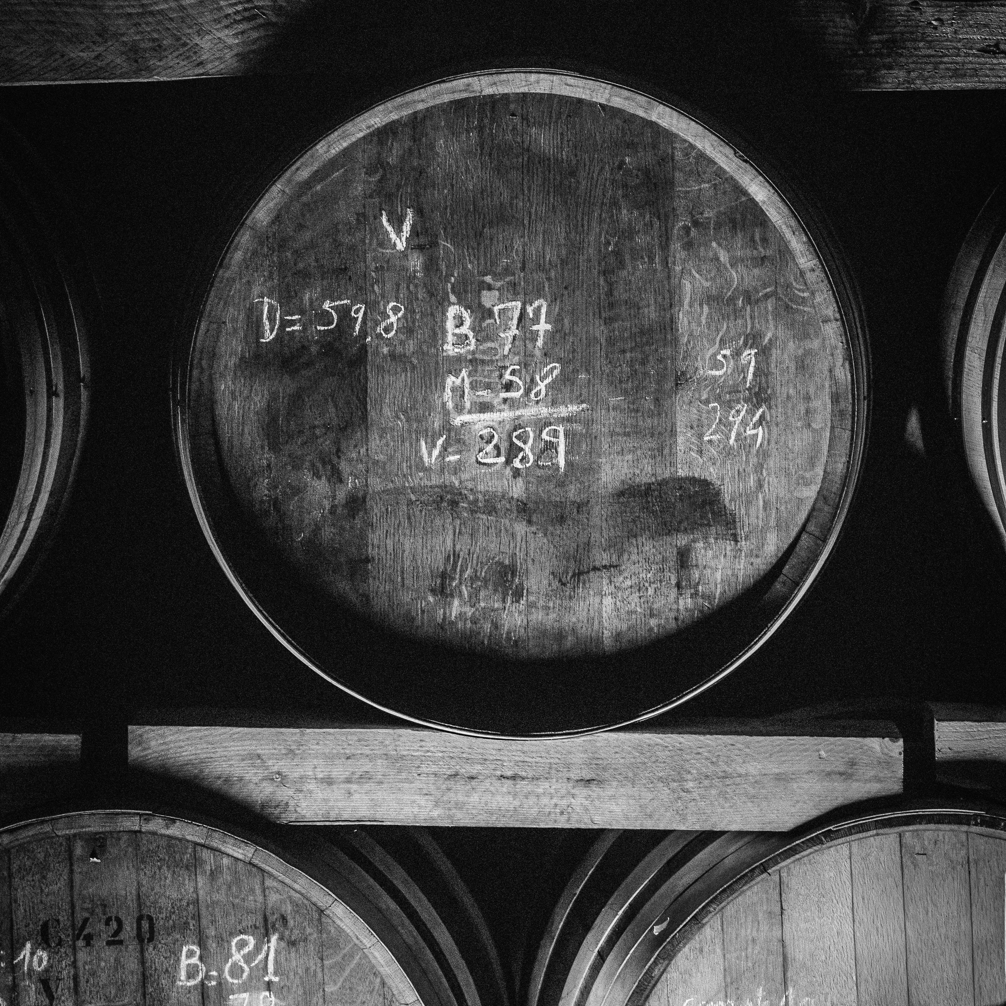 Photographie en noir et blanc d'un fond de fût en bois dans un chai de vieillissement, affichant des inscriptions et codes de traçabilité écrits à la craie blanche - Sainte Harmonie, Baijiu Français.