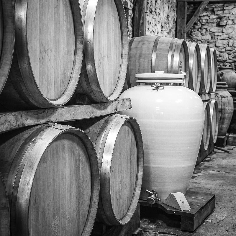 Grande jarre en porcelaine blanche vernie stockée aux côtés de tonneaux en bois pour le vieillissement du spiritueux dans une cave traditionnelle en pierre, photo en noir et blanc - Sainte Harmonie, Baijiu Français.