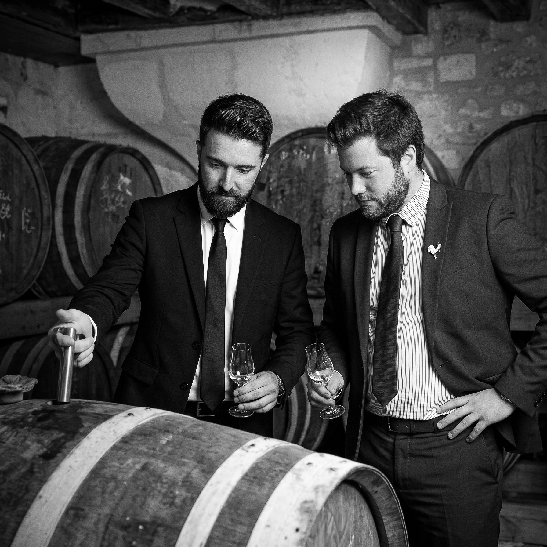 Portrait en noir et blanc des fondateurs Matthieu et David en costume dans leur chai prélevant un échantillon et tenant un verre de dégustation près des fûts de vieillissement - Sainte Harmonie, Baijiu Français.